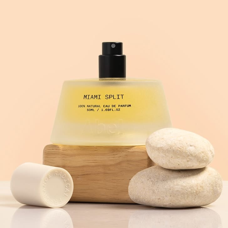 Abel | Miami Split Eau de Parfum | Styled image, frosted glass fragrance bottle with black mister sitting on piece of wood with stones in front of it and a white cap laying beside it against peach background