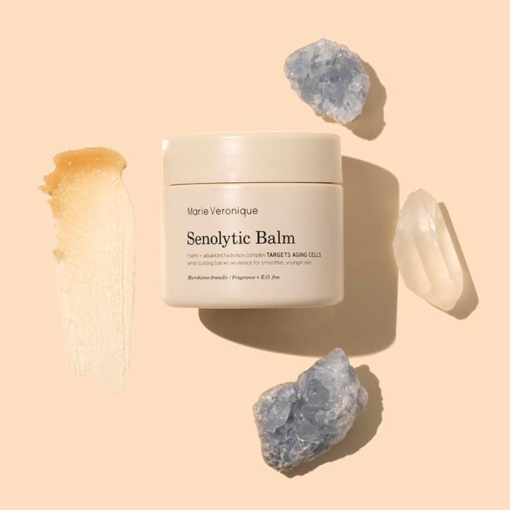 Marie Veronique | Senolytic Balm | Styled Image flat lay with swatch | Beige jar with golden product swatch on peach background with crystals
