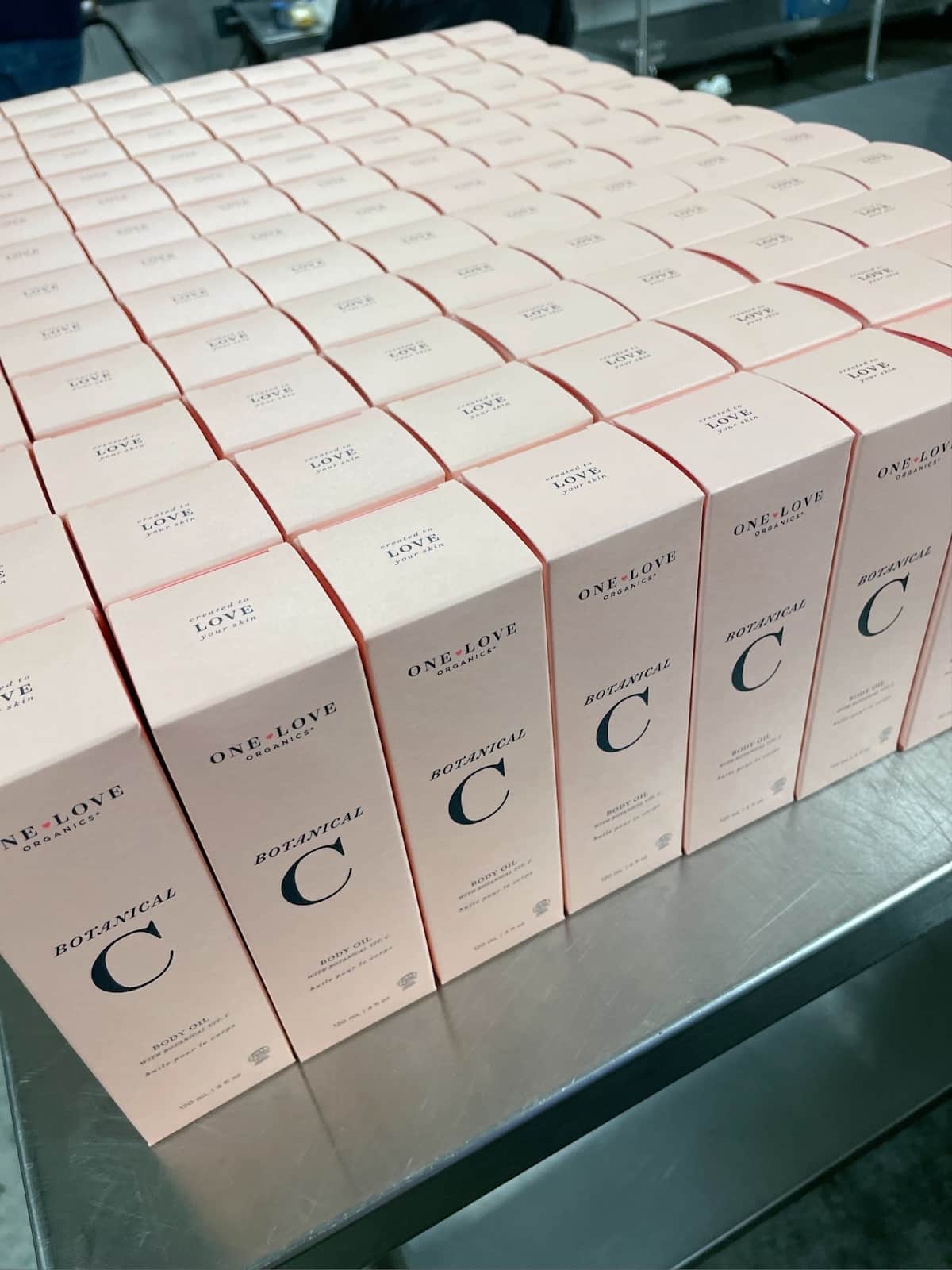 Close-up of One Love Organics Botanical C Body Oil packaging showing neat rows of fresh, blush-toned boxes with branding visible.